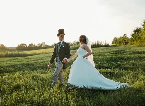 Braut in weißem Kleid und Bräutigam mit Zylinder laufen über eine Wiesen