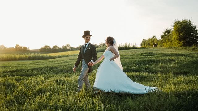 Braut in weißem Kleid und Bräutigam mit Zylinder laufen über eine Wiesen